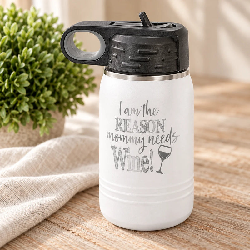 A white insulated water bottle with a black lid and a gray printed slogan reading 'I am the REASON mommy needs Wine!' with a wine glass icon, shown on a wooden surface with a beige cloth and green plant in the background.