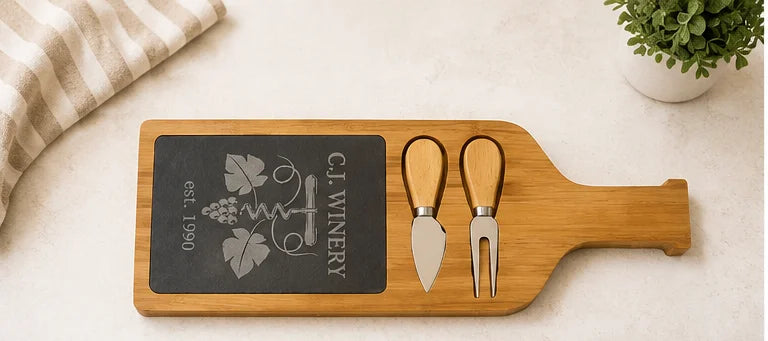 A wine bottle-shaped wooden serving board with a black chalkboard insert engraved with a grapevine and corkscrew design, plus two utensils with light wood handles, styled on a light surface with a folded cloth and small plant in the background.