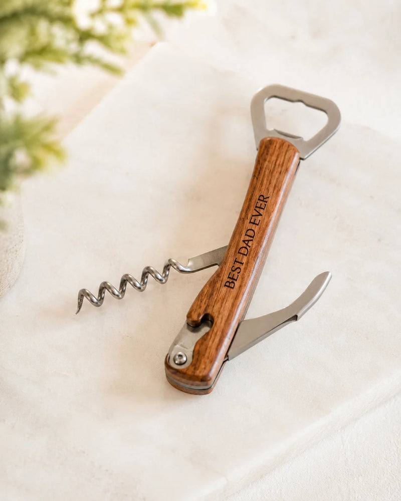 A wooden-handled corkscrew bottle opener with a metal corkscrew and bottle opener bit extended, engraved with the words 'BEST DAD EVER', resting on a light marble surface.