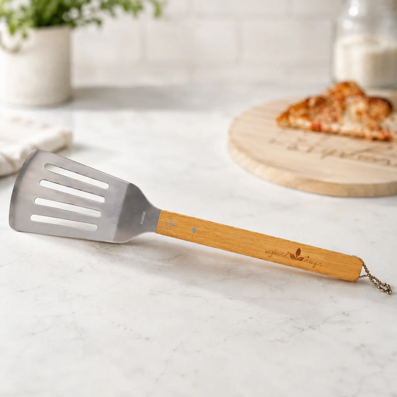 A wooden-handled metal spatula with a small chain loop at the end, resting on a light marble countertop. The background shows a blurred kitchen scene with a pizza on a wooden board and a potted plant.