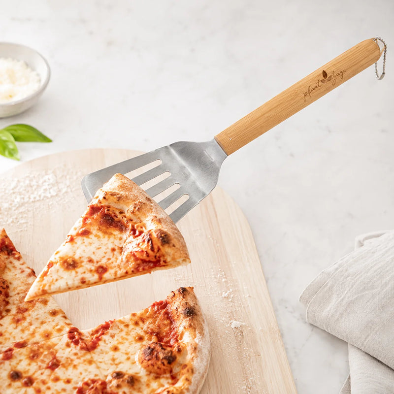 A wooden-handled pizza peel/spatula with a metal blade lifting a slice of cheese pizza on a light wood board, set on a white marble surface with a small bowl of basil and grated cheese in the background.