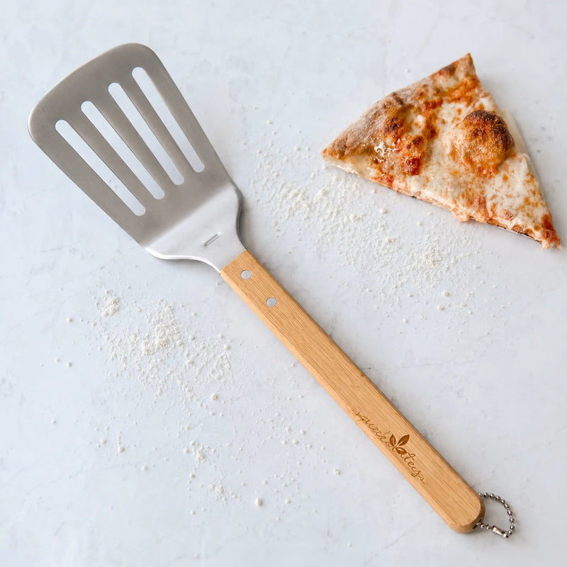 A wooden-handled pizza spatula with a stainless steel slotted head, placed on a light surface with flour scattered around and a slice of pizza nearby. The handle is engraved with the brand name 'Pizzeria Bella'.