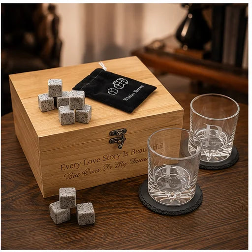 A wooden gift box engraved with 'Every Love Story Is Beautiful But Wines Are My Favorite' sits on a dark wooden table. On the box are several small gray speckled drink cubes and a black velvet pouch with a white logo.