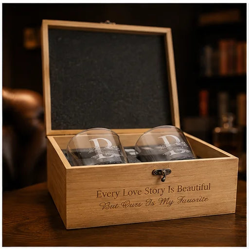 A wooden gift box with an open lid, containing two clear engraved drinking glasses. The front of the box is engraved with the phrase 'Every Love Story Is Beautiful But Yours Is My Favorite' and has a small metal latch on the front.