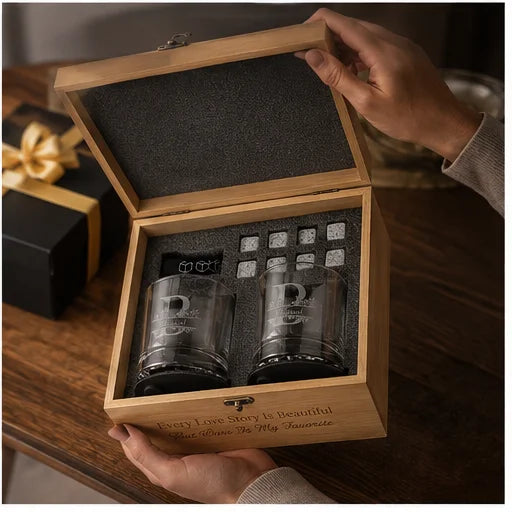A wooden gift box with an open lid is held by hands on a wooden table. Inside are two clear engraved glasses with black bases, surrounded by small square gift tags.
