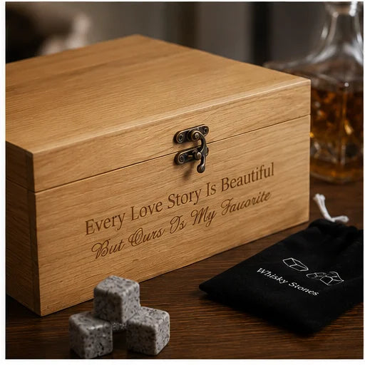 A wooden gift box with engraved text reading 'Every Love Story Is Beautiful But Ours Is My Favorite,' shown on a wooden surface with three gray whiskey stones and a black drawstring pouch labeled 'Whiskey Stones.' A blurred bottle appears in the background.