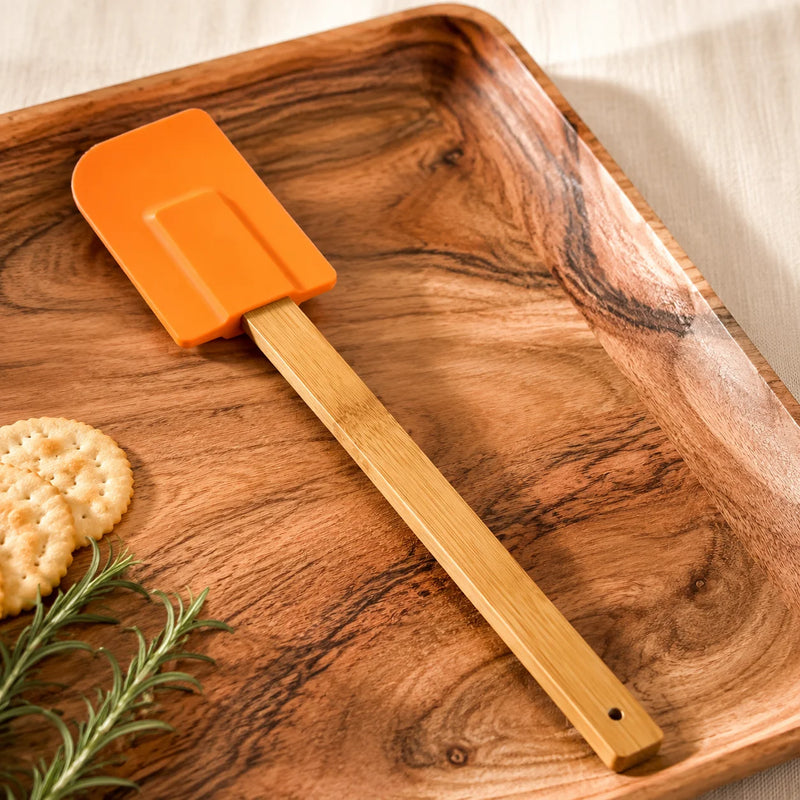 An orange silicone spatula with a light bamboo handle rests diagonally on a polished wooden serving tray, styled with crackers and rosemary.