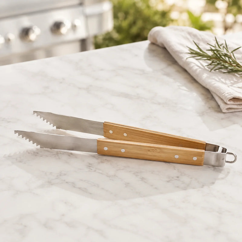 Bamboo-handled kitchen tongs with serrated stainless steel blades, resting on a white marble countertop with a blurred kitchen background.
