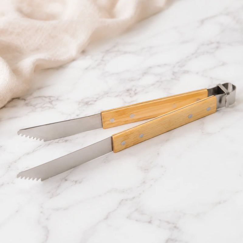Bamboo-handled metal tongs with serrated blades, photographed on a white marble surface with a soft beige cloth in the background.