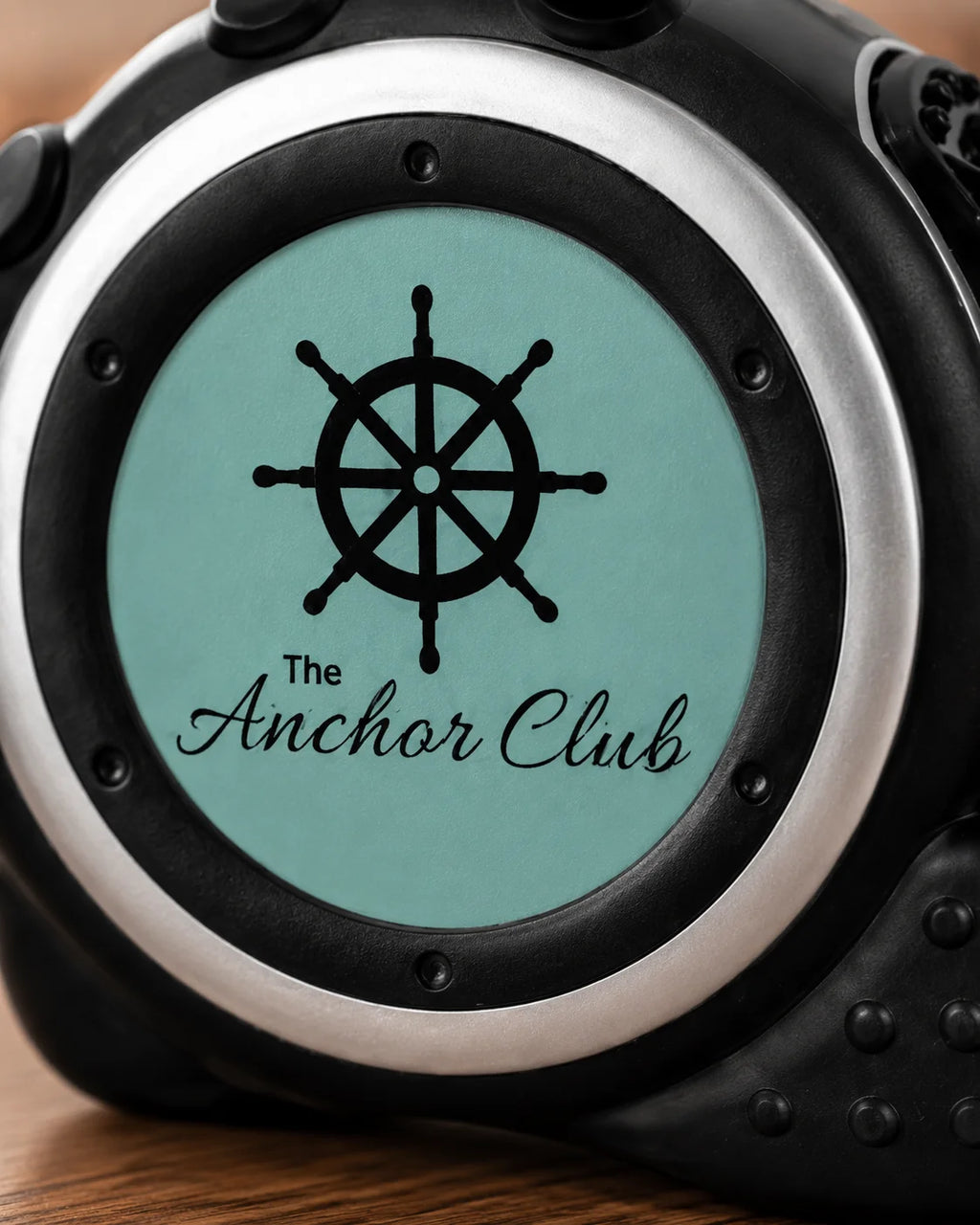 Close-up of a custom watch face with a black ship's wheel graphic and the text 'The Anchor Club' on a light teal background, surrounded by a silver bezel and black casing.
