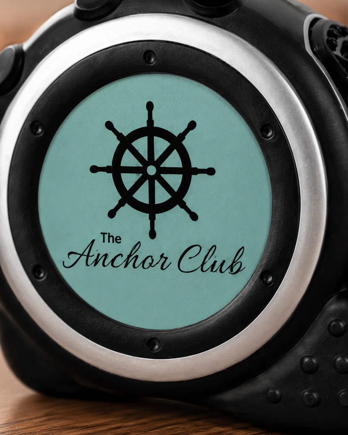 Close-up of a custom watch face with a black ship's wheel graphic and the text 'The Anchor Club' on a light teal background, surrounded by a silver bezel and black casing.