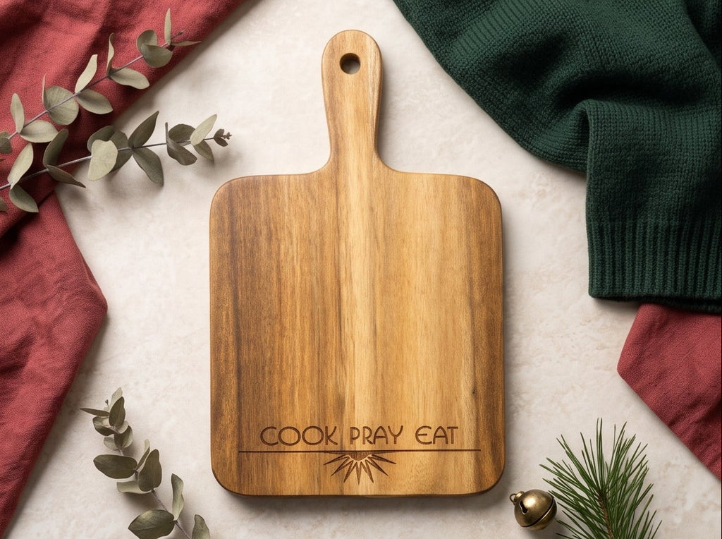 Four-panel collage showing the same wooden cutting/serving board from different angles, styled with eucalyptus leaves, pine branches, a green knit sweater, and red fabric. The board is engraved with the words 'COOK PRAY EAT' and a sunburst motif.
