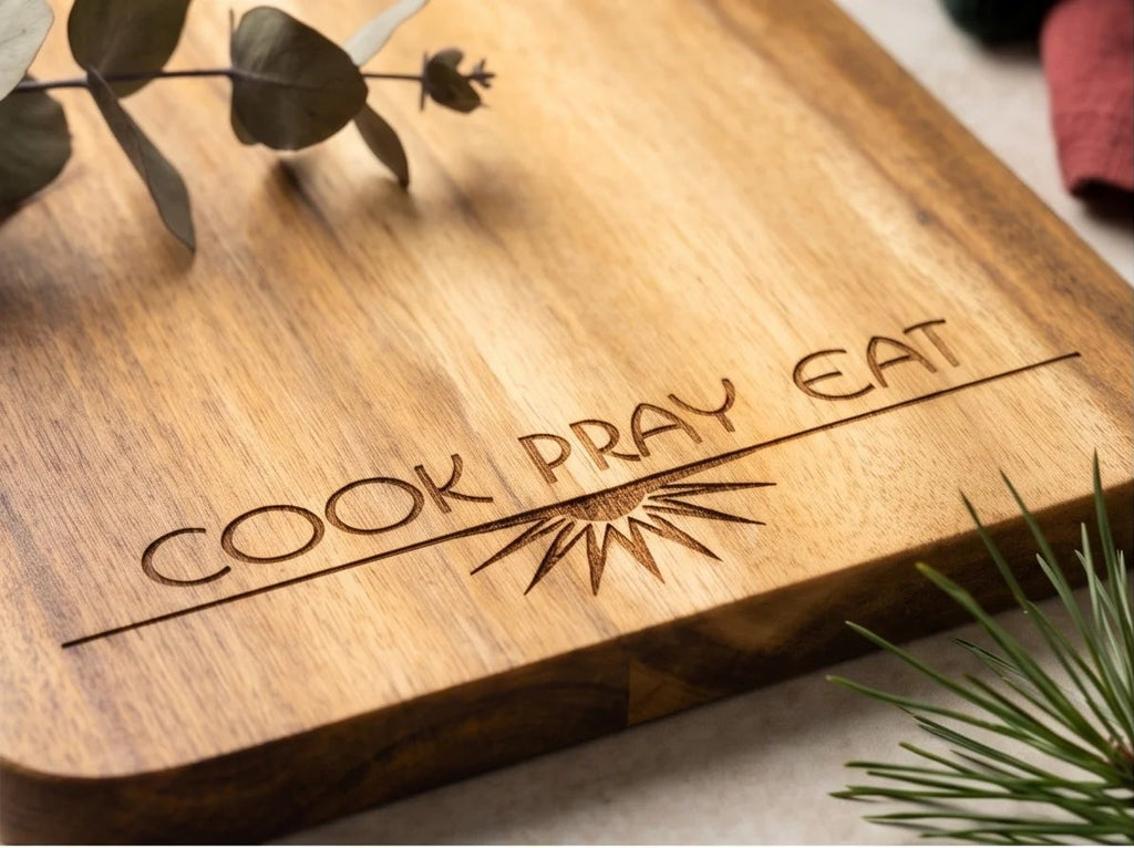 Four views of a rectangular wooden serving board with a handle, engraved with 'COOK PRAY EAT' and a sunburst design. The boards are shown in different angles with holiday styling including green wool, eucalyptus leaves, pine branches, and small gold bells on a light textured surface.