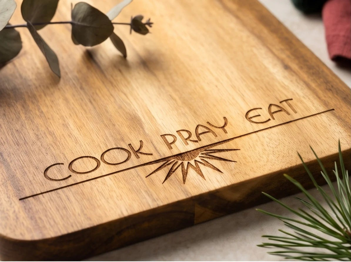 Four views of a rectangular wooden serving board with a handle, engraved with 'COOK PRAY EAT' and a sunburst design. The boards are shown in different angles with holiday styling including green wool, eucalyptus leaves, pine branches, and small gold bells on a light textured surface.