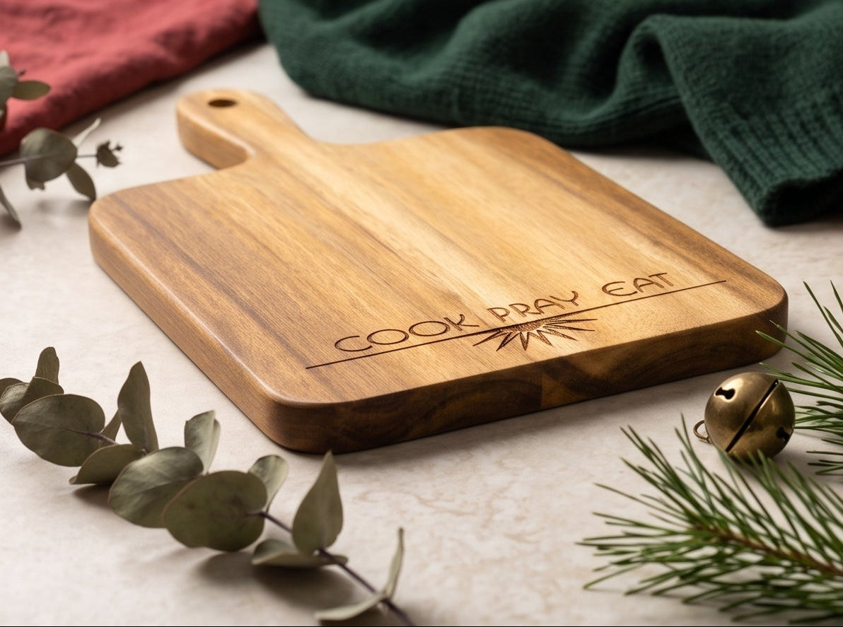 Four views of a rectangular wooden serving board with a handle, engraved with 'COOK PRAY EAT' and a sunburst design. The boards are shown in different angles with holiday styling including green wool, eucalyptus leaves, pine branches, and small gold bells on a light textured surface.