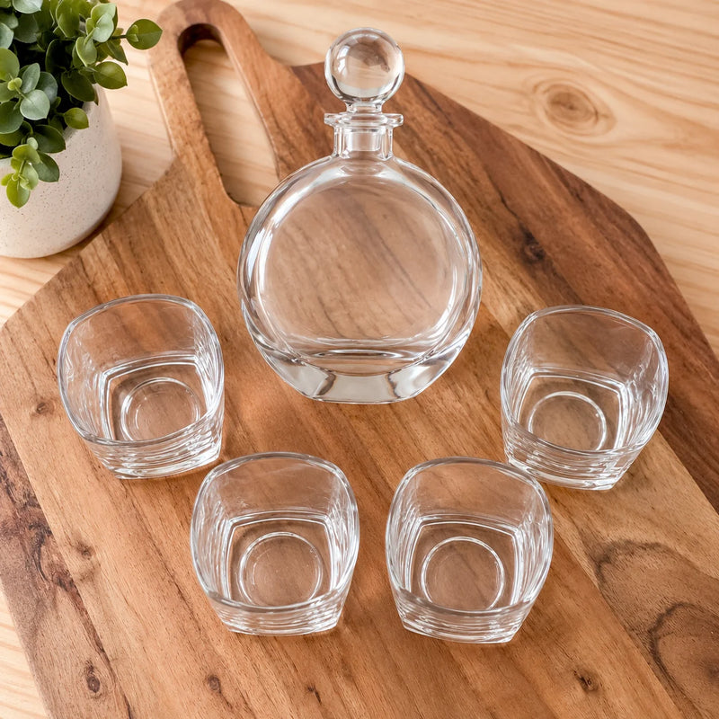 A clear glass decanter with a round stopper and four matching small clear glasses arranged on a wooden serving board, with a potted plant in the background.