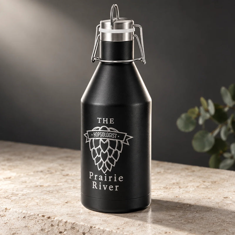 A matte black insulated growler bottle with a metal swing-top lid, placed on draped cream fabric with hops branches and cones as styling props. The bottle is printed with white text reading “THE HOPSLOGIST” and “Prairie River.